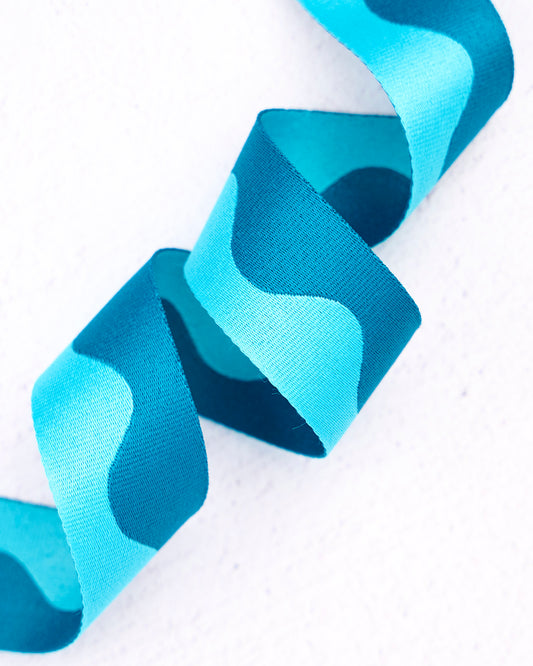 Close-up of wavy nylon webbing by Renaissance Ribbons, featuring a flowing pattern in bright aqua blue and deep teal blue, curled into gentle loops against a clean white background.