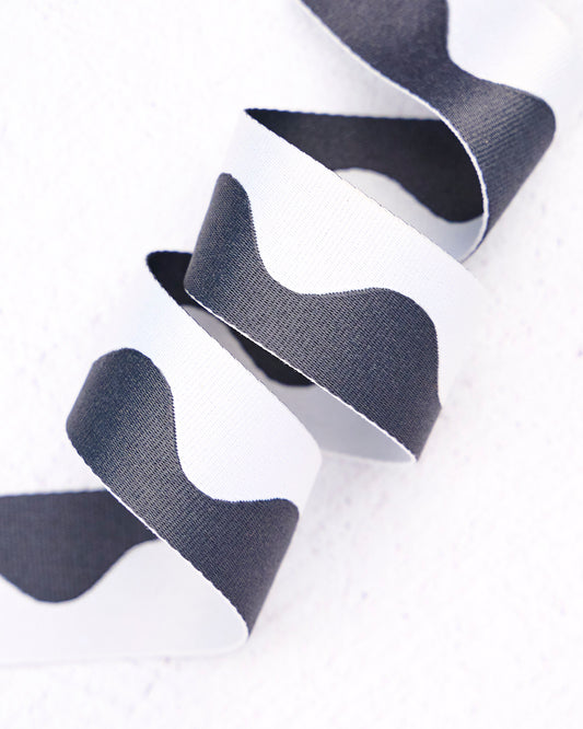 Close-up of wavy nylon webbing by Renaissance Ribbons, featuring a flowing pattern in crisp white and deep charcoal gray, curled into gentle loops against a clean white background.