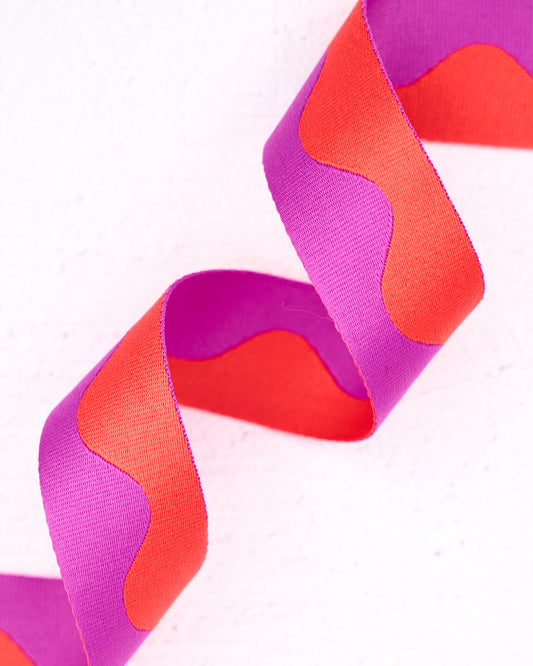 Close-up of wavy nylon webbing by Renaissance Ribbons, featuring a flowing pattern in vivid magenta-purple and bright coral-orange, curled into gentle loops against a clean white background.