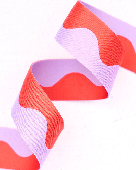 Close-up of wavy nylon webbing by Renaissance Ribbons, featuring a flowing pattern in light lavender (pastel purple) and bright coral-red, curled into soft curves against a clean white background.