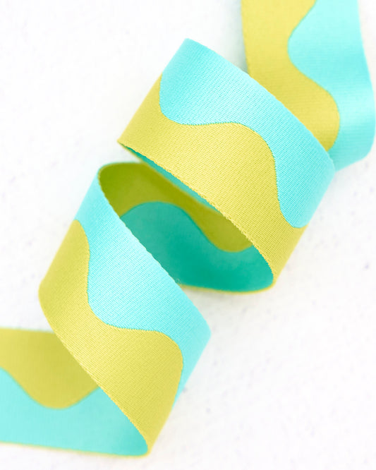 Close-up of wavy nylon webbing by Renaissance Ribbons, featuring a flowing pattern in soft aqua blue and light yellow-green (lime), curled into gentle loops against a clean white background.