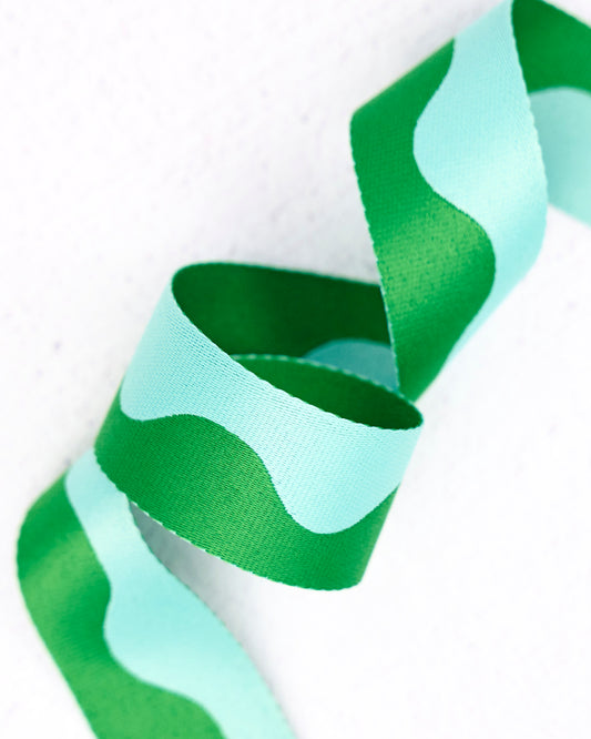 Close-up of wavy nylon webbing by Renaissance Ribbons, featuring a flowing pattern in light mint green and rich emerald green, curled into gentle loops against a clean white background.