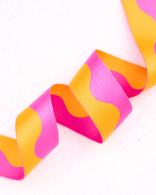 Close-up of wavy nylon webbing by Renaissance Ribbons, featuring a flowing pattern in bright hot pink and vibrant golden orange, curled into gentle loops against a clean white background.