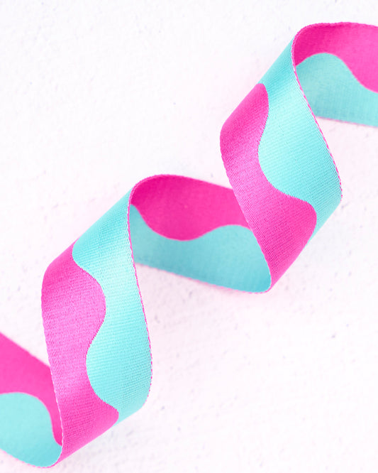 Close-up of wavy nylon webbing by Renaissance Ribbons, featuring a flowing pattern in bright hot pink and soft aqua blue, curled into gentle loops against a clean white background.
